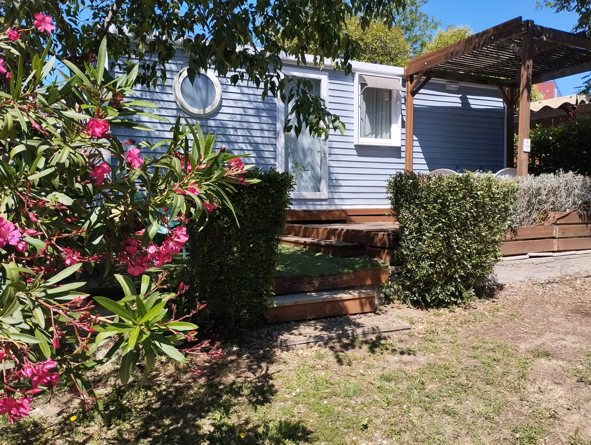Mobil-home bleu avec pergola et buisson de fleurs roses.