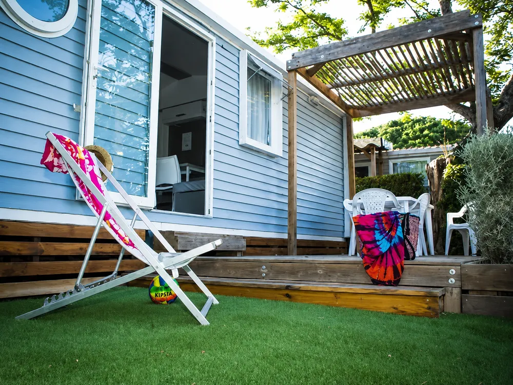 Blue mobile home with wooden terrace, pergola and lounger in the foreground