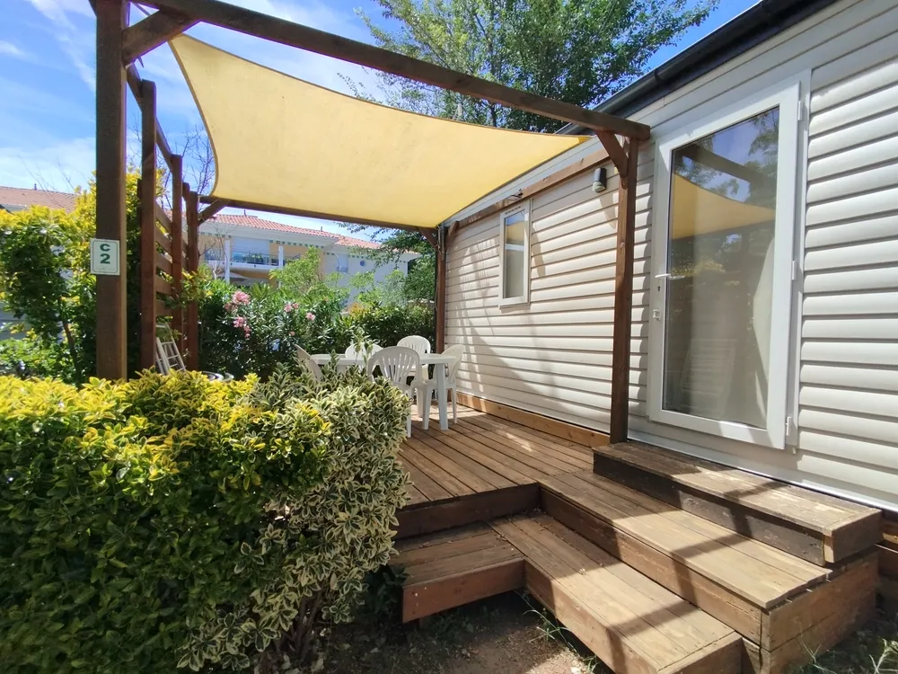 Terrasse en bois d'un mobil-home avec table blanche et voile d'ombrage beige.