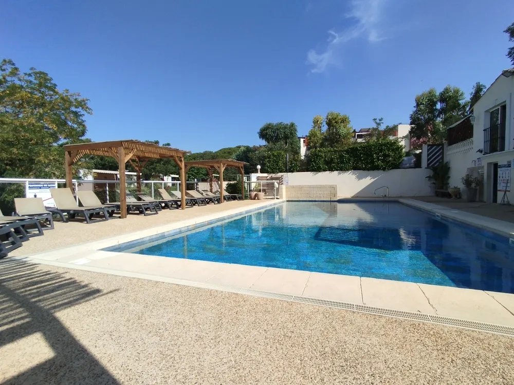 Grande piscine rectangulaire de camping entourée de nombreux transats alignés sous des pergolas en bois par une journée ensoleillée.