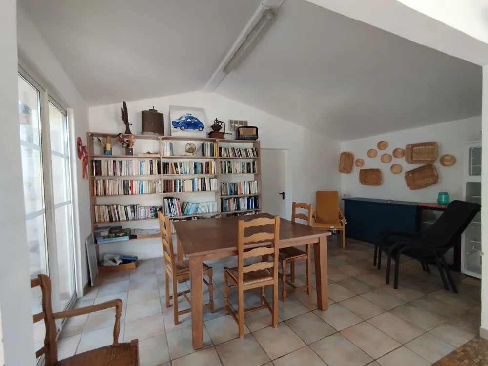 Bright common room featuring a large wooden dining table with straw-bottomed chairs and a large white bookcase filled with books.